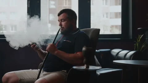 A man sits in front of the windows and smokes a hookah, while looking at his Stock Footage 253410522