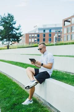 Man sits on grass steps looking at tablet in a city setting during daytime Stock Photos