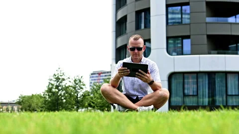 Man sits on grass using a tablet beside a modern office building during the day Stock Footage 348704332