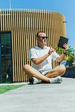 Man sits on the ground and uses a tablet while wearing sunglasses in an out.. Stock Photos