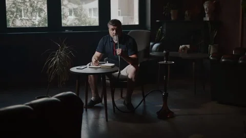 A man sits in a hookah lounge at a table and exhales smoke. Stock Footage 253492296