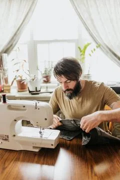 Man sits at kitchen table with sewing machine patching work pants Stock Photos