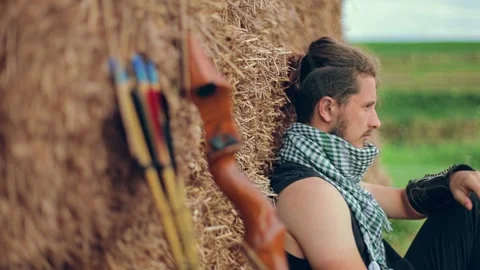 The man sits leaning on sheaves of hay next to him with a bow and arrow Defocus Видео 135592016
