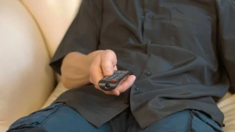 A man sits on a leather sofa, pressing buttons on a black remote control Stock Footage 297815108