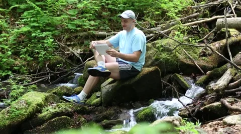Man sits near small river in the forest and communicates via tablet computer 스톡 동영상 59143012