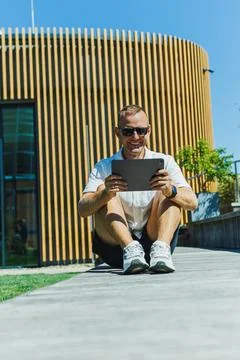 Man sits on pavement using tablet outside modern building with wooden wall .. Stock Photos