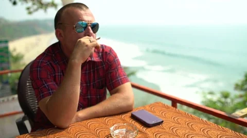A man sits in a restaurant at a table by the sea and smokes a cigarette Stock Footage 74759090