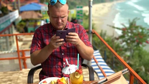 A man sits in a restaurant at a table by the sea and takes pictures on the phone Stock Footage 74761750