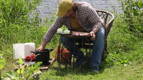 A man sits on the river bank at a table and cooks meat for shish kebab Stock Footage 278538290