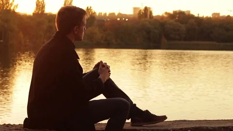 A man sits by the river at sunset and looking thoughtfully into the distance Stock Footage 96388770