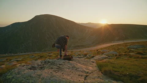 Man sits on rocks watching the sun rise over mountains while a road winds Stock Footage 345634738