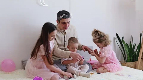 A man sits on a rug while children play with toys. One child paints his nails, Stock Footage 336462729