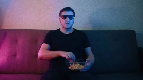 A man sits on a sofa in 3D glasses eating chips. Stock Footage 131376379