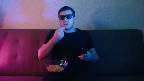 A man sits on a sofa in 3D glasses eating chips and switching channels on a TV. Stock Footage 131822666