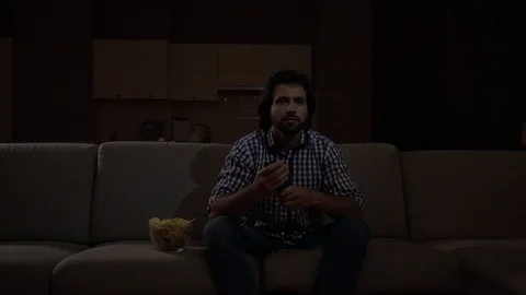 Man sits on sofa and cheers. He watches TV. Guy is worried. He holds remote Stock Footage 99742821