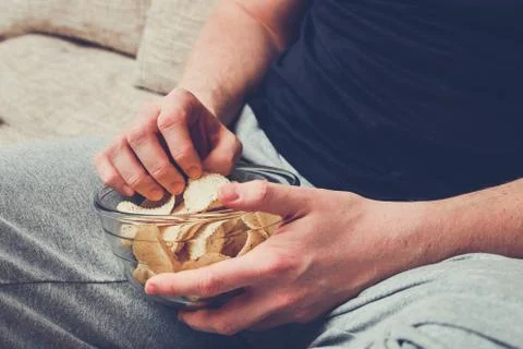 A man sits on a sofa and eats chips from a glass dish. Stock Photos