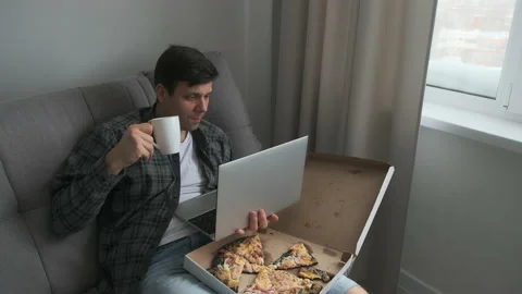 Man sits on sofa eats tasty pizza drinks tea watches in laptop sings a song. Stock Footage 152969450