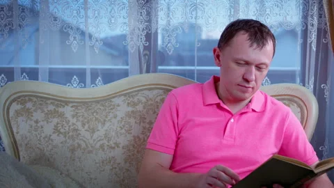 Man sits on a sofa by the window and reads a book with interest, an evening Stock Footage 303762709