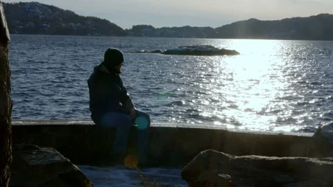 Man sits on stone bench at sunset overlooking ocean, lost in thought, contemplat Stock Footage 233898346