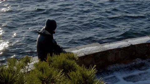 Man sits on stone bench at sunset overlooking ocean, lost in thought, contemplat Stock Footage 233898364