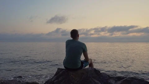 A man sits at sunset on a large stone and relaxes. Rear view silhouette of a man Stock Footage 117710344