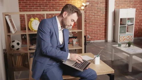 A man sits on a table and types into a laptop, the laptop lies on his leg. Vidéo 194363346