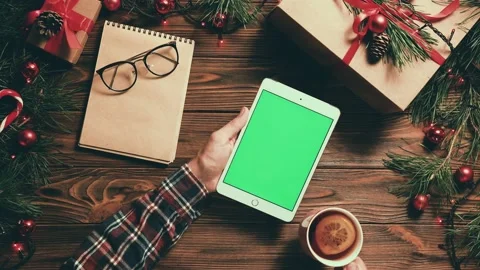 Man sits at table with Christmas decor and reads e-book on tablet while drinking Stock Footage 143746417