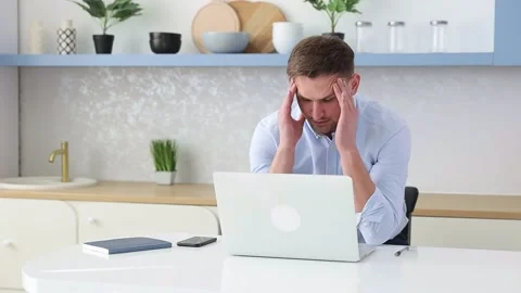 A man sits at a table with a laptop and holds his head. Irregular working day Stock Footage 152904109