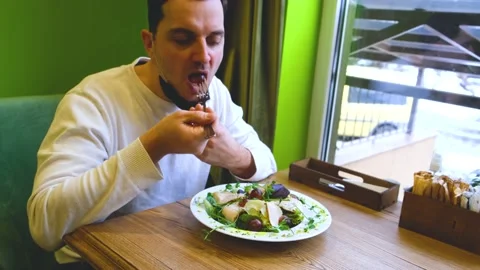 A man sits at a table in a restaurant wearing a mask, lowers the mask puts food Stock Footage 169961946