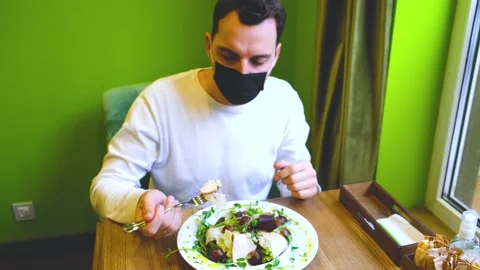 A man sits at a table in a restaurant wearing a mask, lowers the mask puts food Stock Footage 169961992