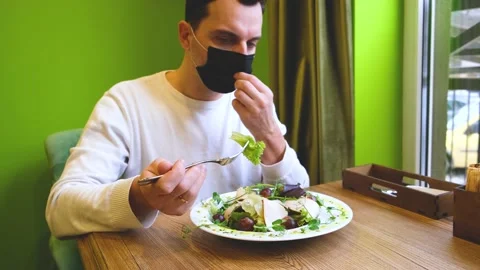 A man sits at a table in a restaurant wearing a mask, lowers the mask puts food Stock Footage 169962042