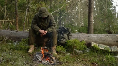 The man sits on the trunk of a fallen tree in a forest camp by the campfire 스톡 동영상 239141210