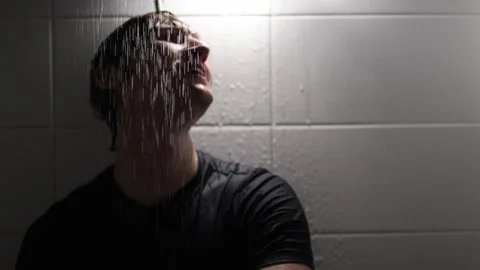 Man sits under shower water smiling with wet hair and dark shirt illuminated by  Stock Footage 315991870