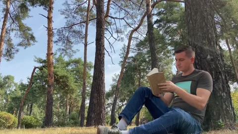 A man sits under a tree and reads a book. The boy gains knowledge in the forest Stock Footage 283554969