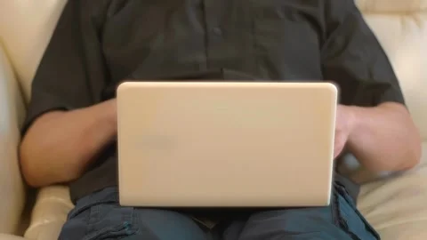 A man sits on a white leather sofa and works absorbedly on a white laptop Stock Footage 297744758