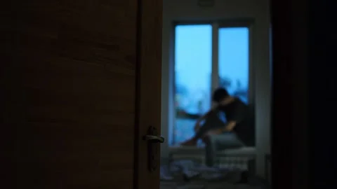 A man sits on a windowsill in a dark room, viewed through an open door, expressi Stock-Footage 296882992