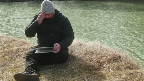 A man sits on the winter grass and works on a laptop-transformer on the bank Vidéo 170305324