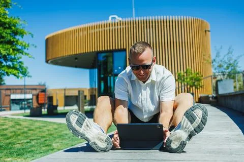 Man sits on a wooden bench using a tablet outdoors near a modern building o.. Stock Photos