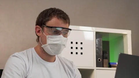 A man sits working at a laptop in a protective mask and goggles Stock Footage 129049302