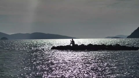 A man sitting alone on the rocks by the sea taking photos Stock Footage 289543004