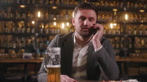 Man is sitting at bar table, engaged in a phone conversation while a half-full Stock Footage 284030873