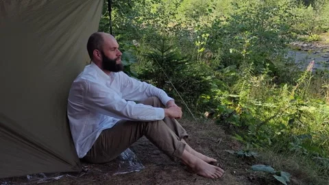 Man sitting barefoot outside camping tarp shelter in pine forest Stock Footage 344887935