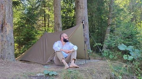 Man sitting barefoot outside camping tarp shelter in pine forest Vídeo Stock 344894131