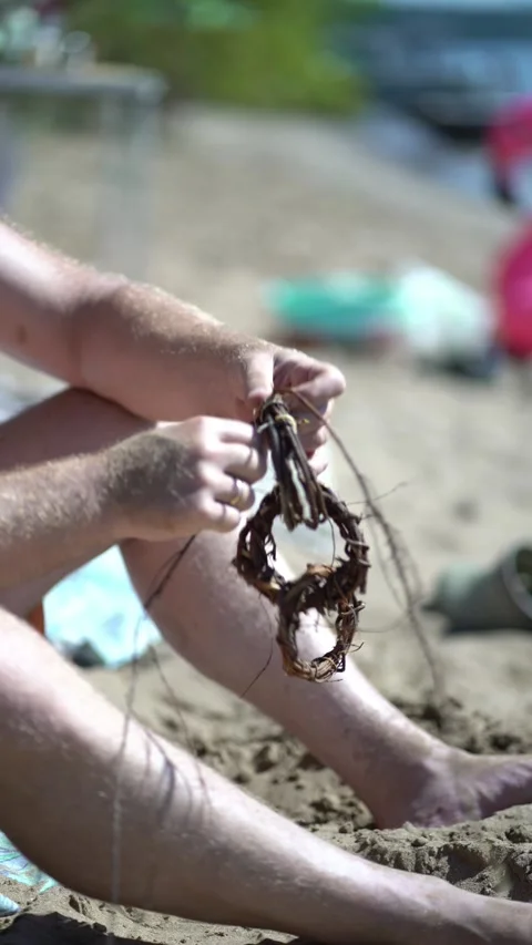 Man sitting barefoot on a sunny beach carefully weaves dry roots and vines Stock Footage 342083844