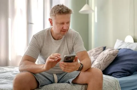 Man sitting on a bed checking his phone during the morning Stock Photos