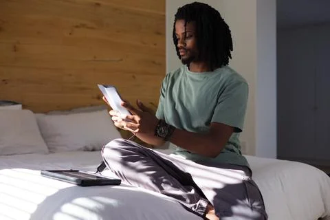 Man sitting on bed cross-legged wearing sage shirt holding lit tablet, charging Stock Photos