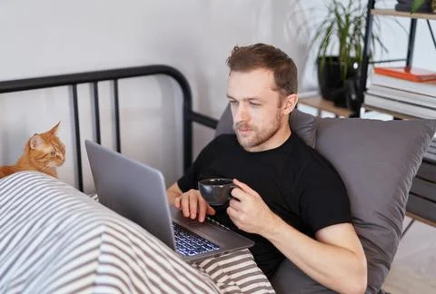 Man sitting on bed with cup of tea using laptop Stock-Fotos