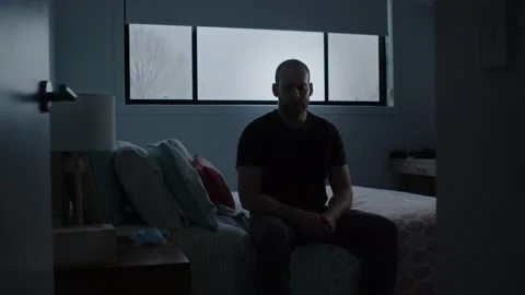 Man Sitting on Bed in Dark Room Puts Face Mask on. Wide shot Pushes In Stock Footage 130576798