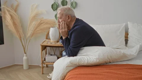 Man sitting on bed with hands covering face, wearing sweater beside nightst.. Stock Footage 339199704