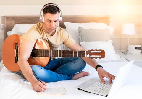 Man sitting on the bed learning to play the guitar. Concept of online lessons Stock Photos